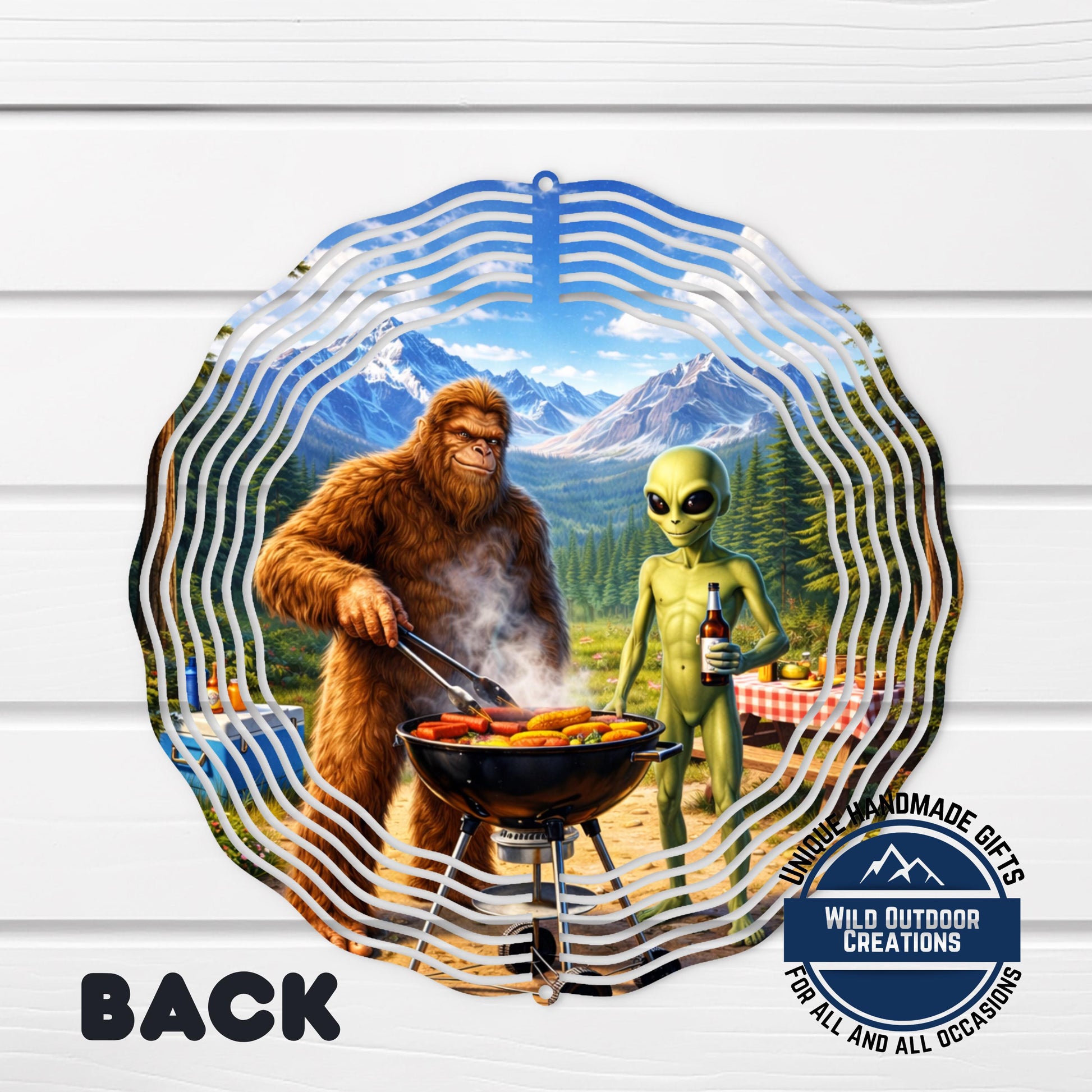 A large metal sculpture depicting a giant brown furry creature cooking food on a grill, with a smaller alien-like creature standing nearby. The sculpture is set against a backdrop of mountains and a clear blue sky.