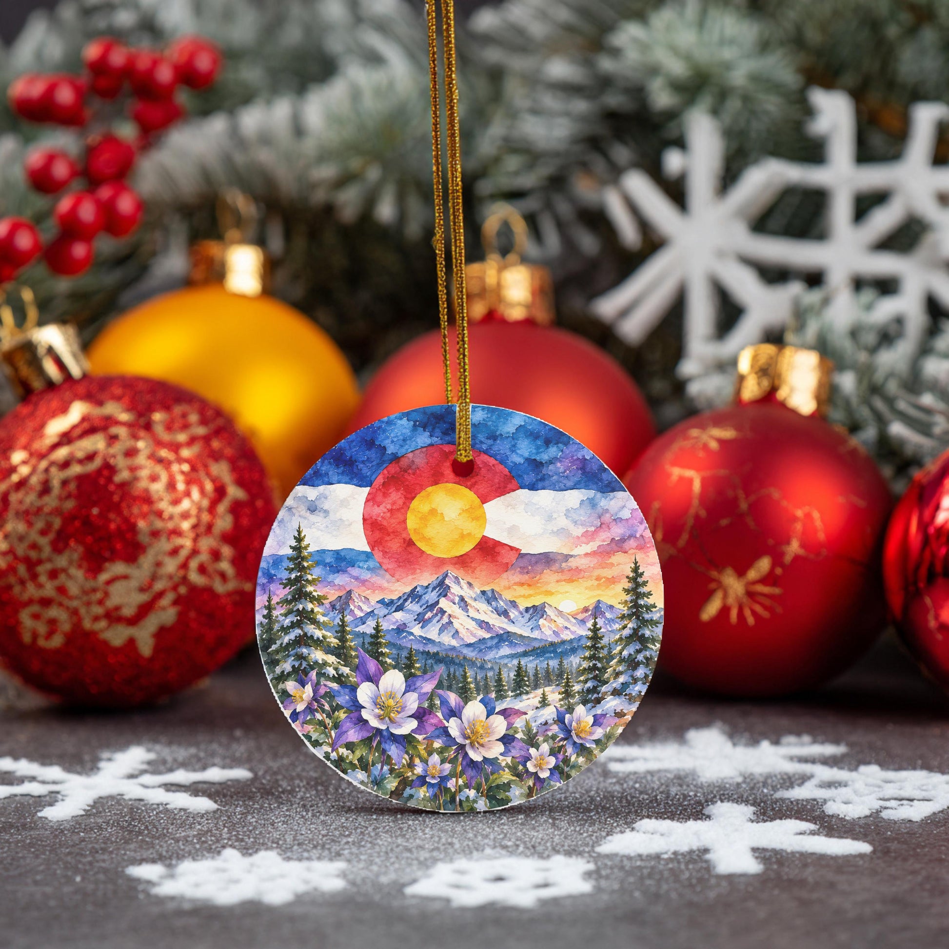A colorful ornament featuring a mountain landscape with a sun in the sky, surrounded by snowflakes and pine trees, is displayed among other Christmas ornaments and decorations.