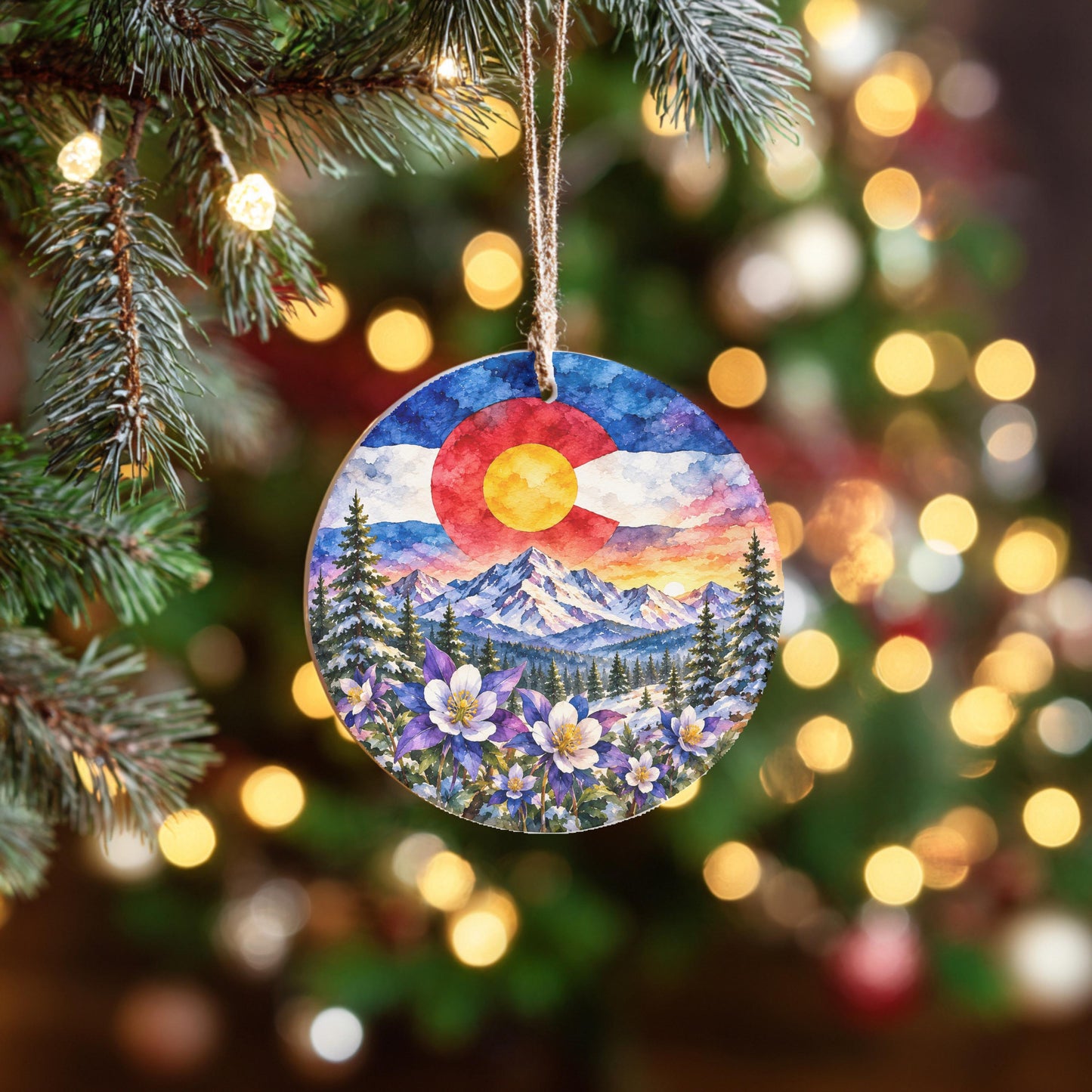A colorful ornament featuring a mountain landscape with a sun in the sky, surrounded by pine trees and flowers, hanging on a Christmas tree.