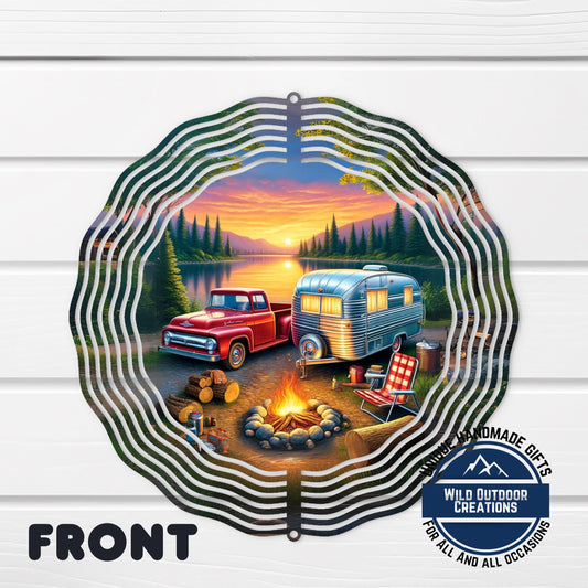 A metal wind chime with a camping scene depicted on it, featuring a red truck, a silver camper, a campfire, and a lake with trees in the background.